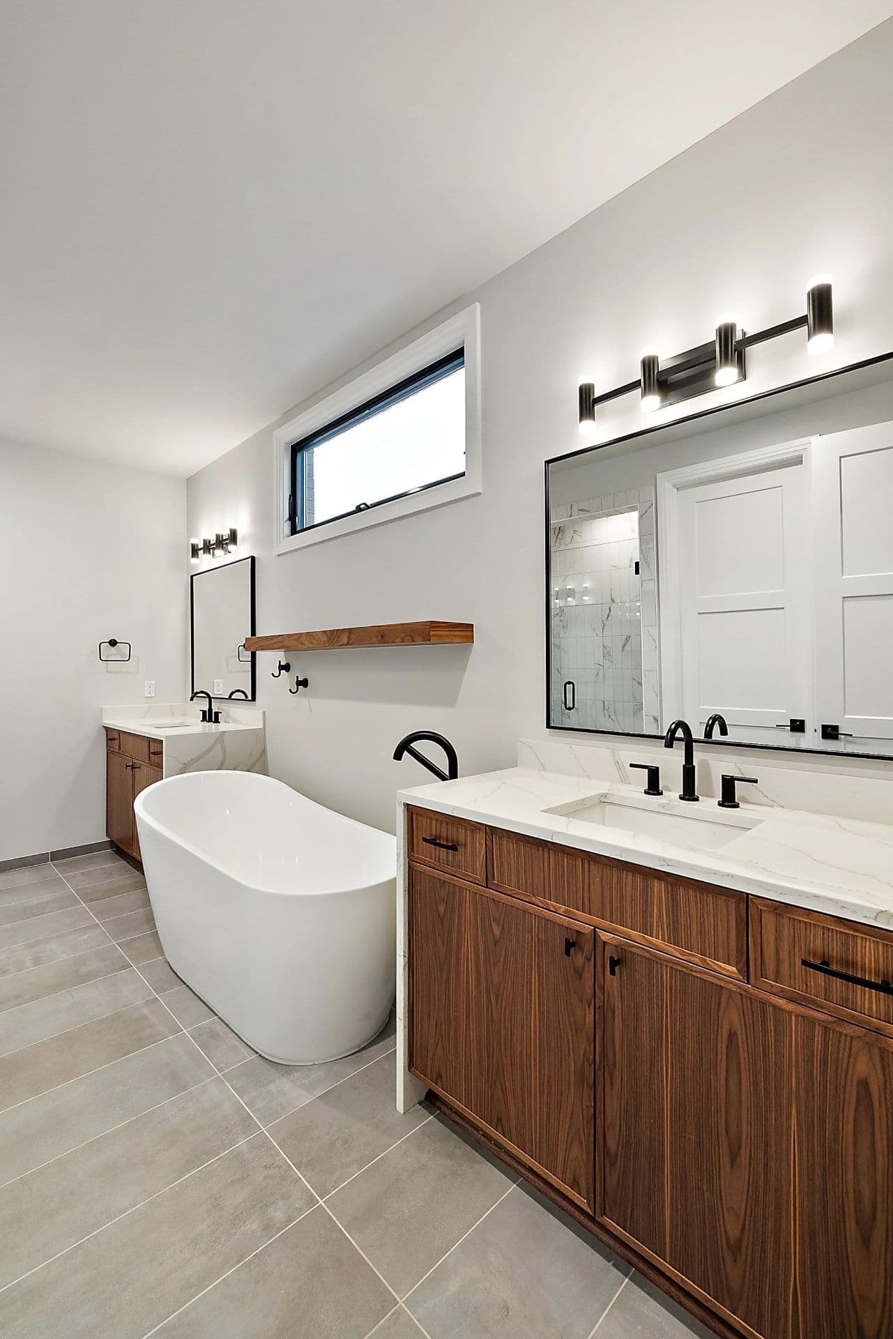 Bathroom Remodeling Design Inspiration Gallery. A white bathroom with large soaking bathtub, double vanity with brown woodgrain cabinets, white marbled countertops, and matte black fixtures.