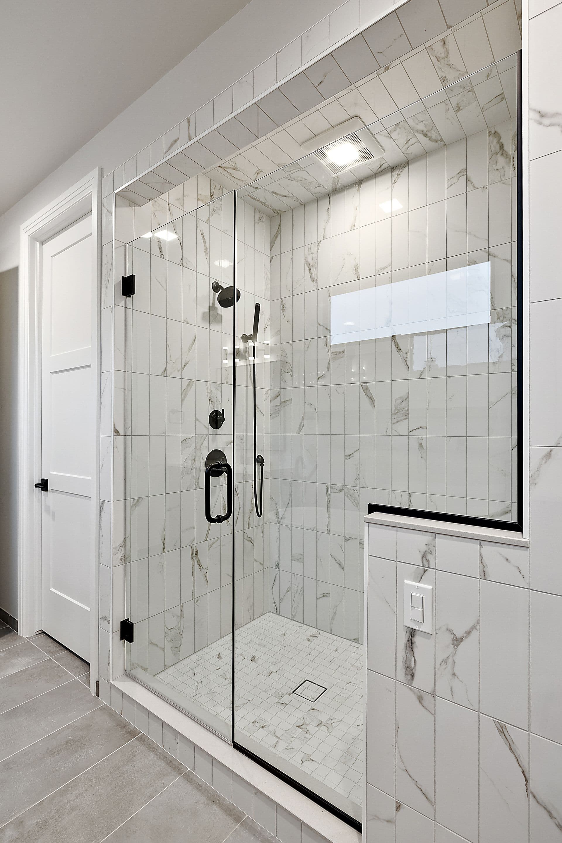 Walk-in shower with floor-to-ceiling white marble-look tile, a frameless glass enclosure with matte black hardware, and a recessed ceiling light.
