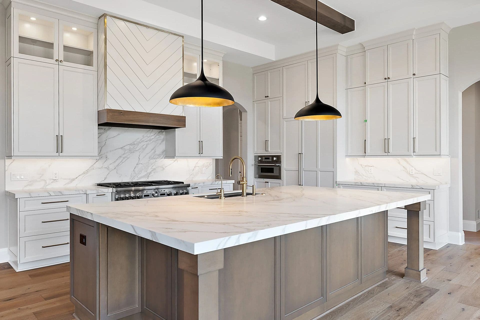 A newly-remodeled kitchen with white cabinets, large central island with white marbled countertop and medium brown cabinets, and black hanging lights.