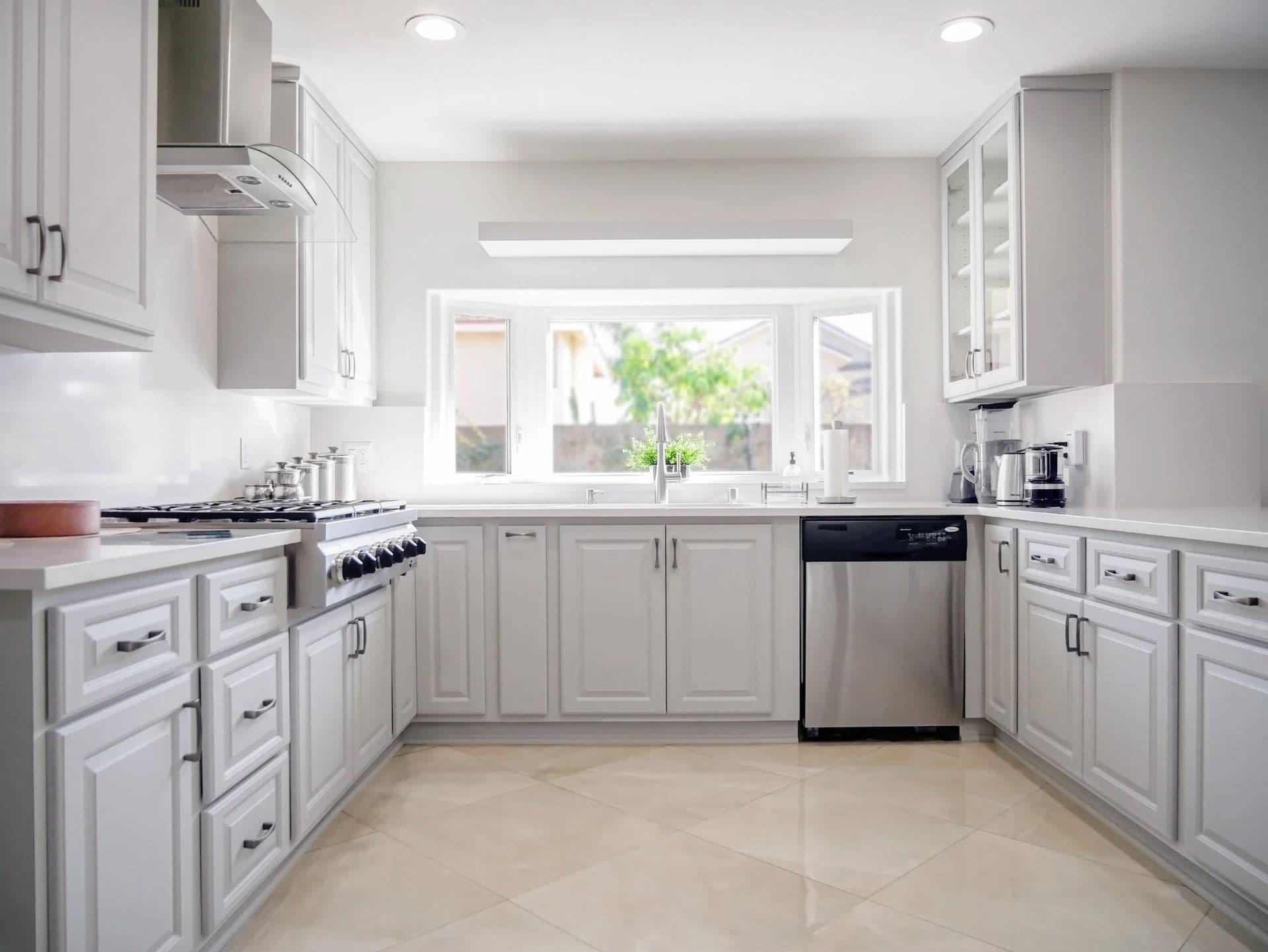 A newly-remodeled white kitchen with off-white cabinets, a large window over the sink, and beige tile floor.