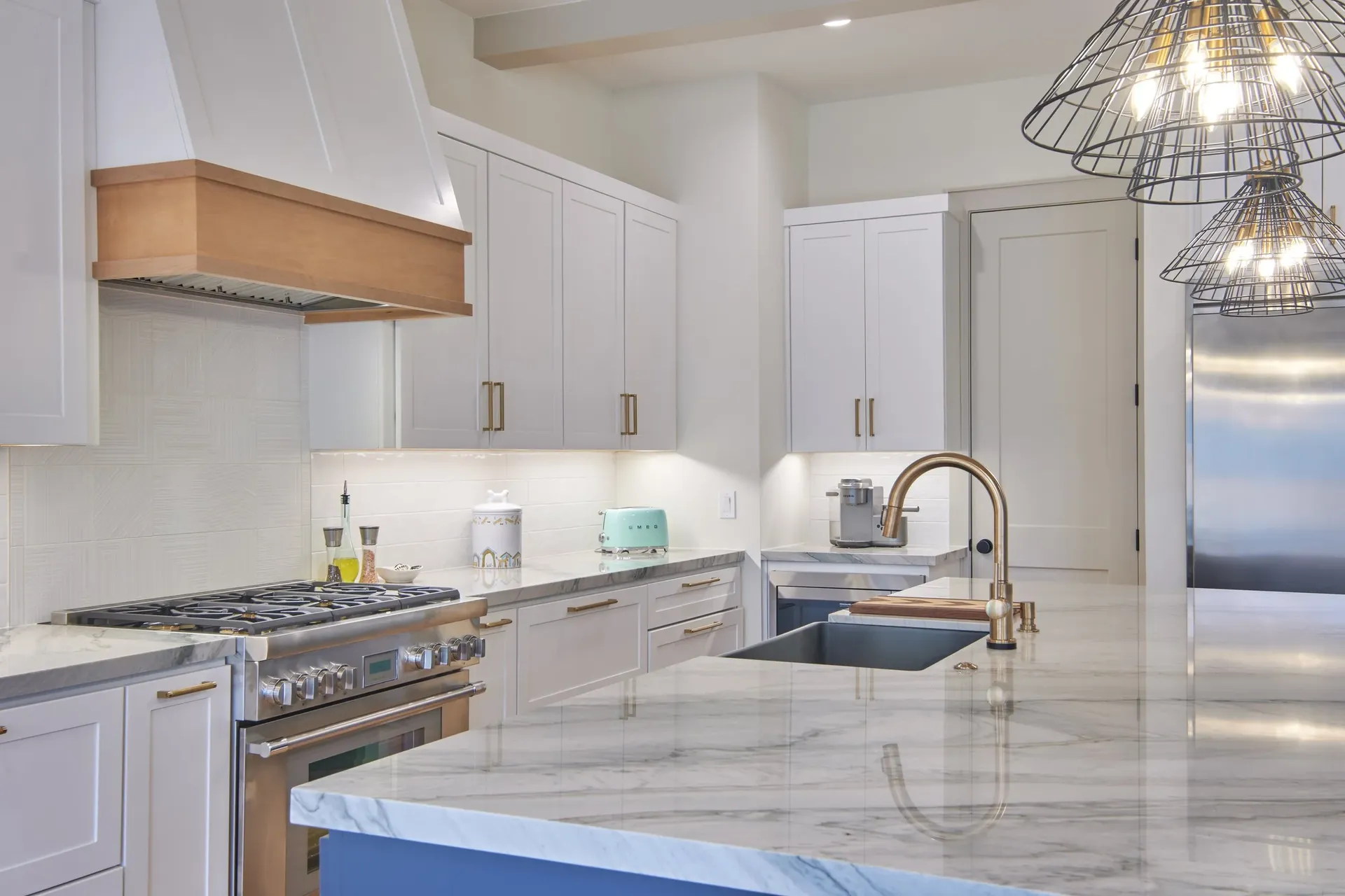 A kitchen with white cabinets and gold fixtures, white tile backsplash, white and natural wood stove hood, and large central island with white and gray marbled countertops.