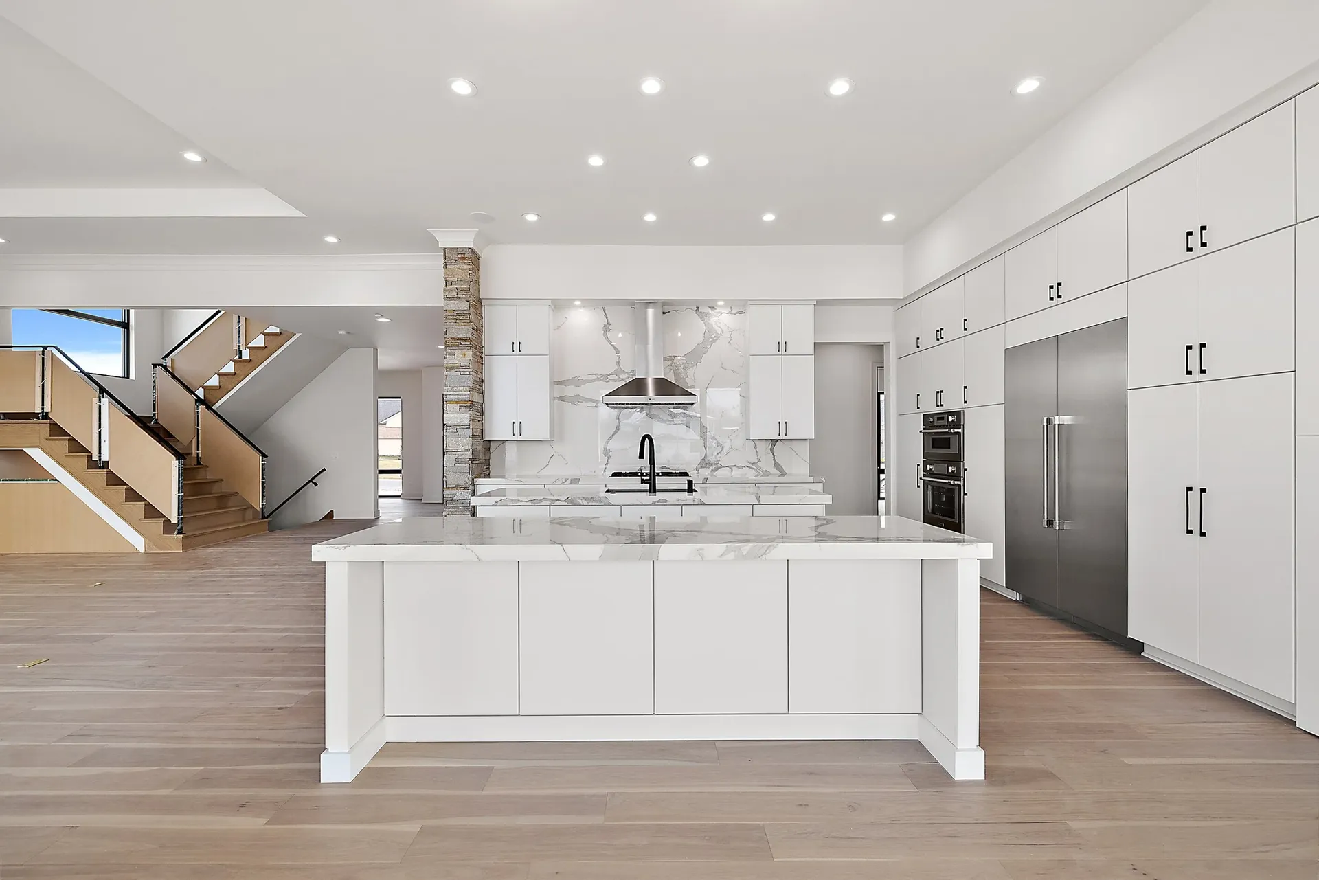 A kitchen with white cabinets, white and gray marbled backsplash wall and countertops, a large central island, and large double door stainless steel fridge.